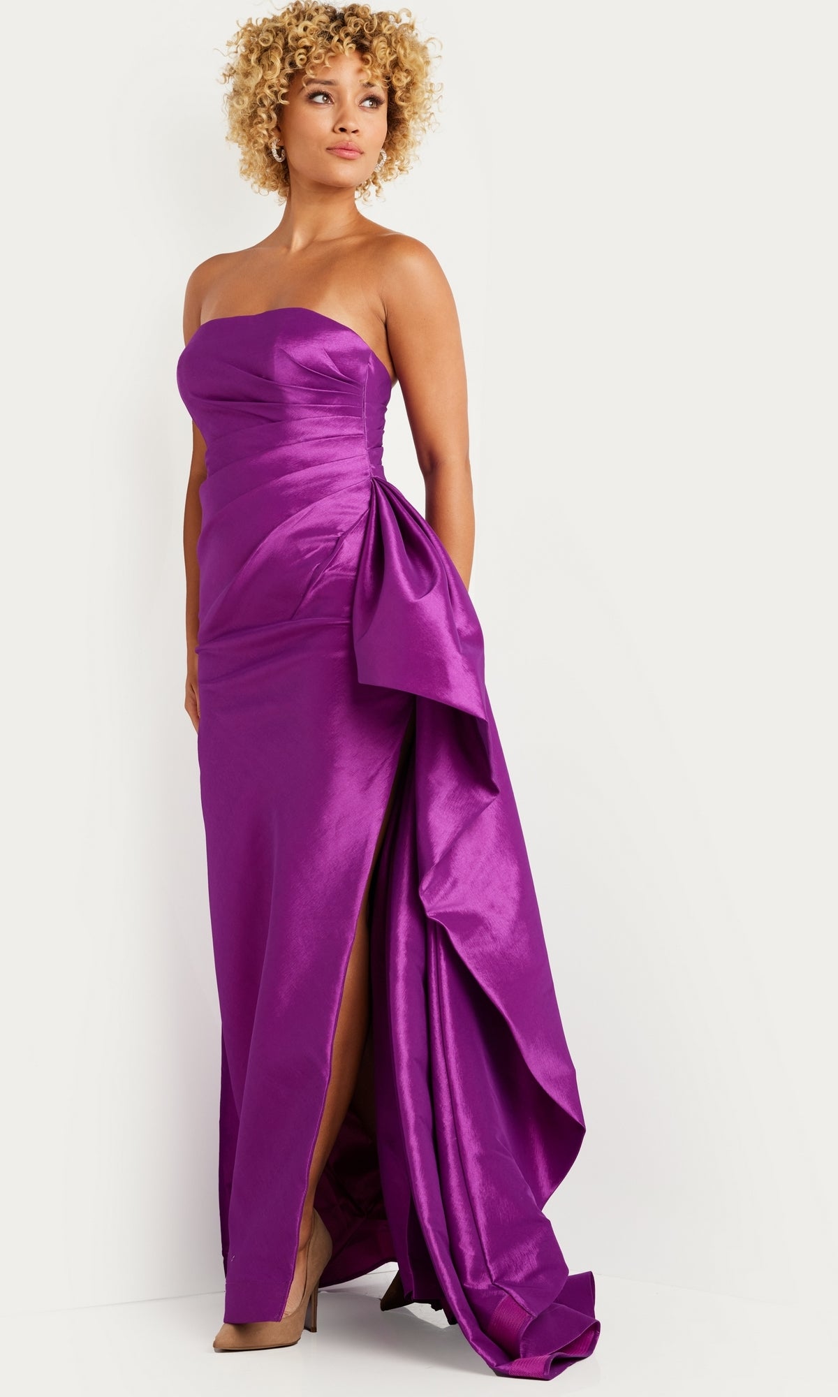 Wearing Jovani’s Long Prom Dress 36854—a bright purple strapless taffeta gown with a side slit and large bow—this person stands against a neutral background, exuding elegant and bold style.